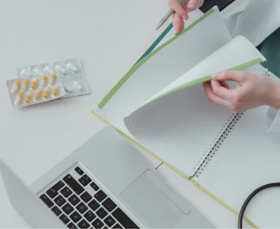 A person is flipping through pages in a notebook with a pen in hand, next to a laptop on a desk.