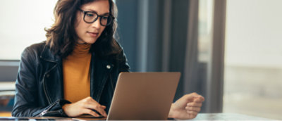 A person wearing glasses and a yellow top under a black jacket is seated at a table, using a laptop 