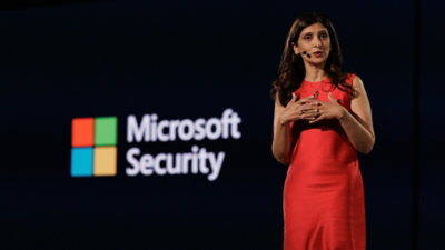 Presenter in a red outfit speaking on stage with Microsoft Security displayed on a screen in the background.