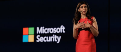 Presenter in a red outfit standing beside a large Microsoft Security logo on a dark background.