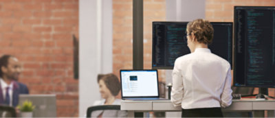 一个人站在桌子旁，在办公室环境中使用笔记本电脑，桌上有多个显示代码的显示器。