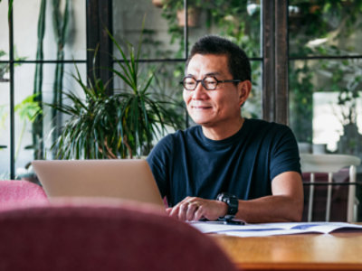 A person wearing glasses and a black shirt sits at a desk, smiling while looking at a laptop. 
