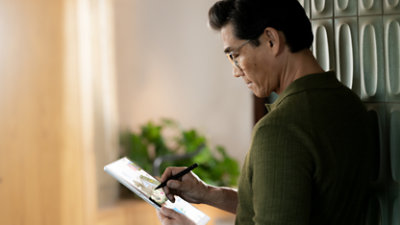 A person wearing glasses uses a stylus to navigate a digital tablet while standing indoors near a plant.