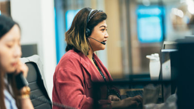 Two people are working at desks in an office, wearing headsets and looking at computer screens. 