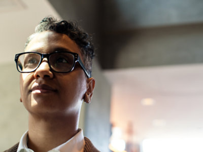 A person with short, curly hair and glasses looks to the side in a well-lit indoor setting.