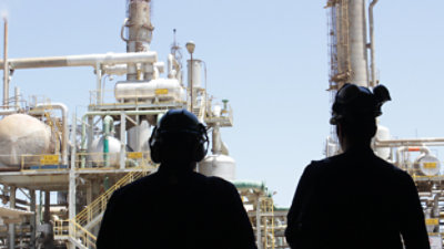 Two industrial workers, silhouetted and wearing helmets, are standing in front of a large refinery structure with pipes 