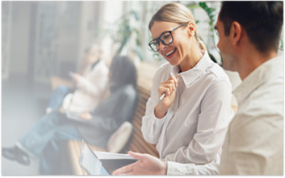 Two people are sitting on a bench indoors, engaged in a conversation and looking at a tablet. 