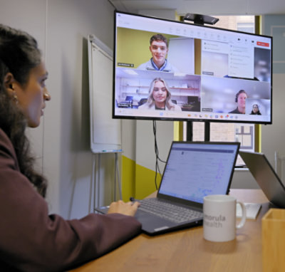 Two people in a meeting room with laptops looking at a television screen displaying four people on a Teams call