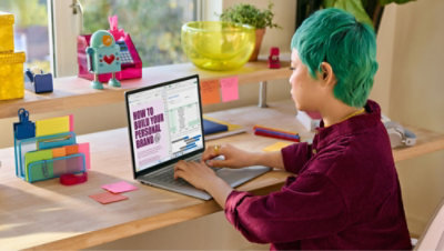 Person with green hair types on a laptop at a desk with colorful decor.