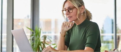 Una mujer con gafas trabaja en un portátil en un escritorio, con una planta y una ventana en el fondo.