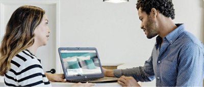 Dos personas miran una pantalla de portátil que muestra una imagen de una cama con ropa en un entorno de habitación.