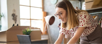 Una mujer con gafas y una camiseta blanca con corazones rojos trabaja en un portátil en una habitación con cajas de cartón y estanterías.