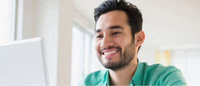 Una persona con cabello oscuro y barba, vestida con una camisa verde, sonríe mientras mira una pantalla de computadora.