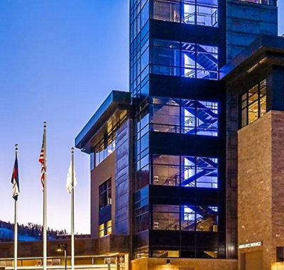 Modern healthcare facility exterior with glass tower, illuminated stairwell, and flagpoles at entrance.