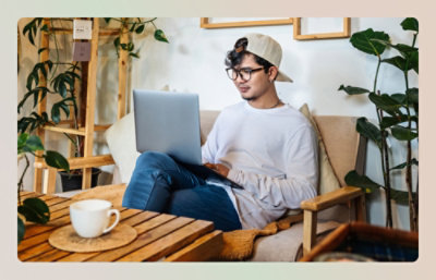 A person uses a laptop while sitting in a room with plants and wooden furniture.