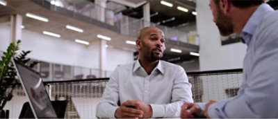 To mænd sidder ved et bord i et kontormiljø og fører en samtale.