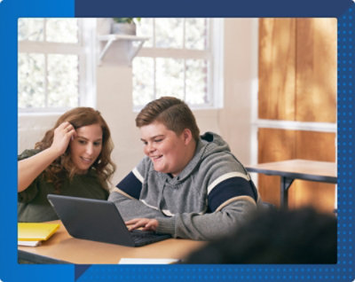 Zwei Personen in einem Unterrichtsraum, die zusammen an einem Tisch sitzen und auf einen Laptop schauen