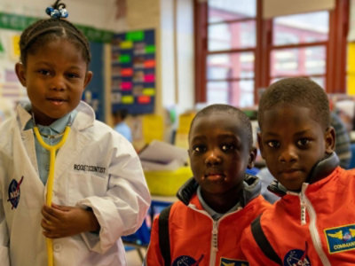 Drei Kinder in einem Klassenzimmer: Eines ist als Wissenschaftler mit einem NASA-Laborkittel verkleidet, die anderen beiden mit Astronautenanzügen.