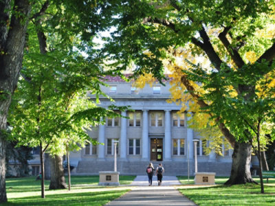 緑と黄色の木々に囲まれた歴史ある大学の建物。入口へと続く道を歩く二人の姿が、静謐なアカデミックな雰囲気を醸し出している。