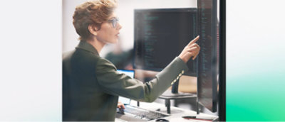 Pessoa de óculos aponta para um código em uma grande tela de computador, de pé, trabalhando em uma mesa com vários monitores.