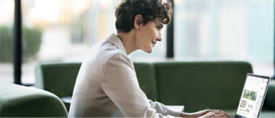 Pessoa de blazer branco sentada em um sofá verde trabalhando em um laptop.