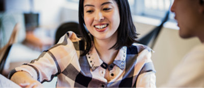 Uma mulher de camisa xadrez sorri enquanto conversa com outra pessoa em um ambiente fechado.