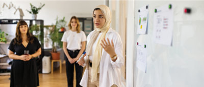 Uma mulher fazendo uma apresentação usando um quadro branco, com três colegas ouvindo. 