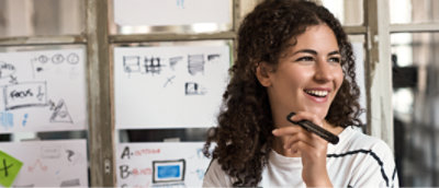 Uma pessoa sorrindo e segurando um marcador está em frente a uma janela com vários diagramas desenhados à mão.