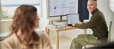 Duas pessoas estão em um escritório bem iluminado. Um está sentado em uma mesa com várias telas exibindo dados e código