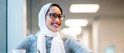 Uma mulher usando óculos e um lenço branco na cabeça está sorrindo sentada perto de uma janela.