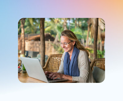 Uma pessoa de óculos sorri enquanto digita em um laptop, sentada em um ambiente externo com árvores e móveis de bambu.