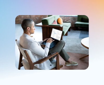 Um homem sentado em uma cadeira usando um laptop em uma sala de estar moderna e ensolarada com sofás verdes e uma parede de tijolos.