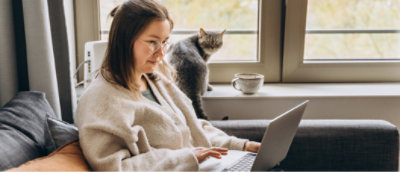 A woman with brown hair wearing glasses sits in front of a laptop with a cat in the background.