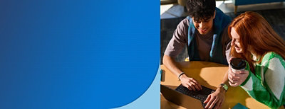 Two students working together at a table on a laptop, one holding a coffee cup.