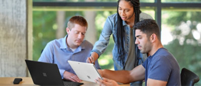 Three people working together, looking at laptop and tablet in a bright room.