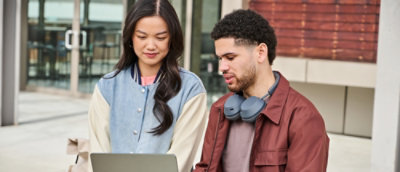 Two people sitting outside using a laptop together near a building entrance.