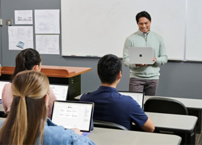 Instructor using laptop while teaching students seated with laptops in a classroom.