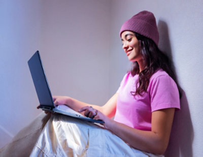 Smiling woman sitting while using a laptop rested on her knees.