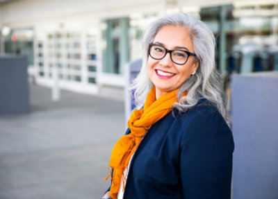Smiling woman with gray hair, glasses, and an orange scarf outdoors.