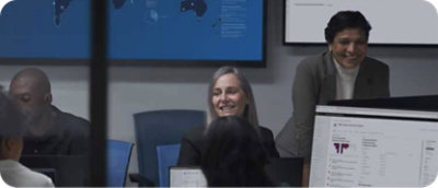 People in a conference room are working on computers with data displayed on screens in the background.