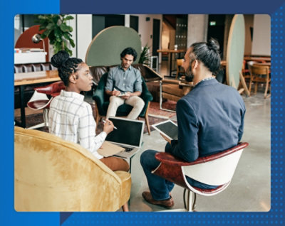 Three coworkers discuss in a modern office; a woman types on a laptop while two men use tablets.