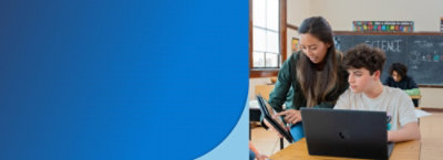 A woman shows something on a tablet to a seated student with a laptop in a classroom.