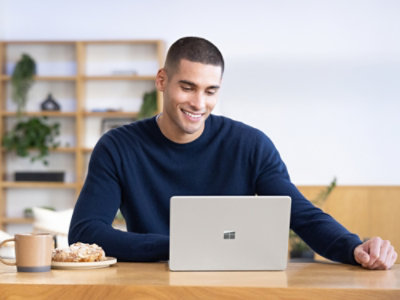 Person using a laptop at a table with a coffee and pastry in a bright home setting.