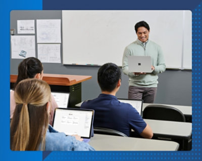 Teacher using a laptop while presenting to students seated in a classroom.