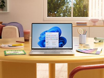 A laptop displaying a colorful home screen is on a round wooden table with various stationery items.