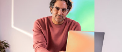 A man in a red shirt and glasses smiling while sitting at a laptop.