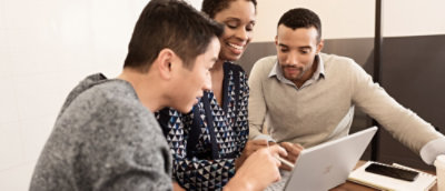 Three people are gathered around a laptop in a modern office setting, engaged in discussion.