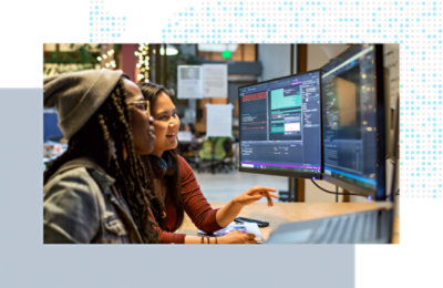 Two coworkers reviewing code together on dual monitors in a shared workspace.