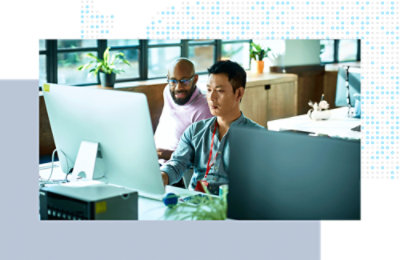 Two coworkers seated at a desk, working together on large desktop monitors in an office.
