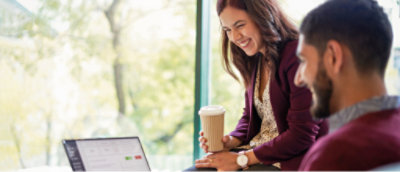 Birisi elinde kupa tutan ve gülümseyen diğeri bir dizüstü bilgisayar ekranına bakan birlikte oturan iki kişi. Arka planda parlak pencere görünümü.